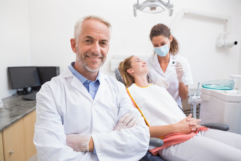 Dentist smiling at camera with assistant and patient behind at the dental clinic